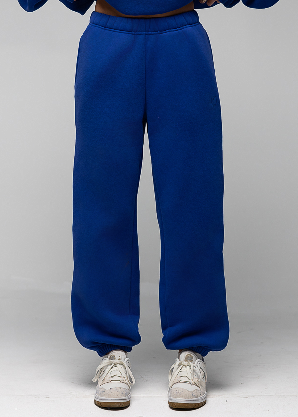 A person wearing a pair of loose-fit royal blue sweatpants with a matching sweatshirt and white sneakers, shown from the chest down against a light gray studio background.