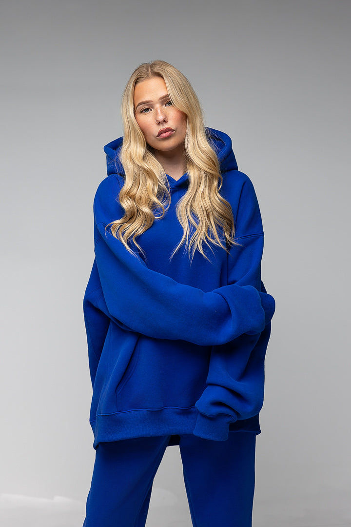 A blonde woman modeling an oversized royal blue hoodie and matching sweatpants against a neutral gray studio background.