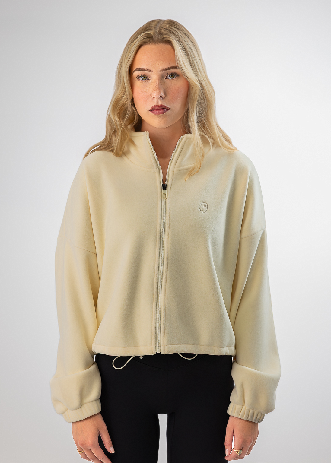 A blonde woman modeling a cream-colored zip-up fleece jacket with a small embroidered logo on the left chest, paired with black pants, against a light gray studio background.