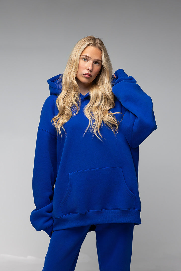 A blonde woman modeling an oversized royal blue hoodie and matching sweatpants against a neutral gray studio background.