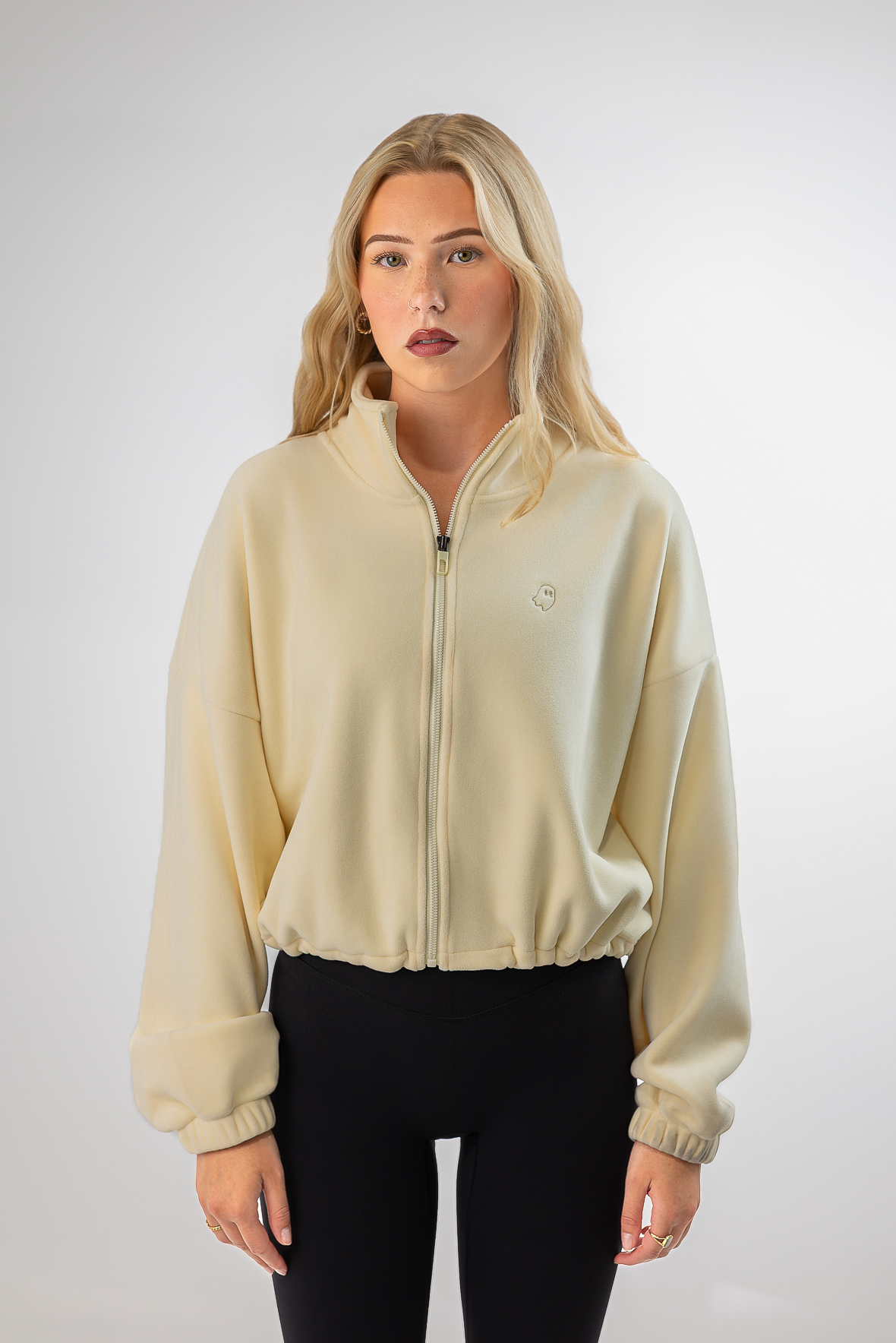 A blonde woman modeling a cream-colored zip-up jacket with a small circular logo on the left chest, paired with black pants, against a light gray studio background.