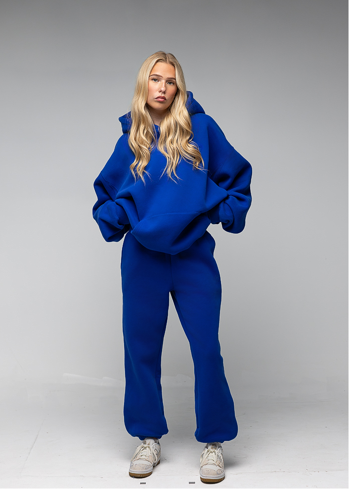 A blonde model wearing a matching royal blue sweatshirt and sweatpants set, paired with light gray sneakers, standing against a neutral gray studio background.