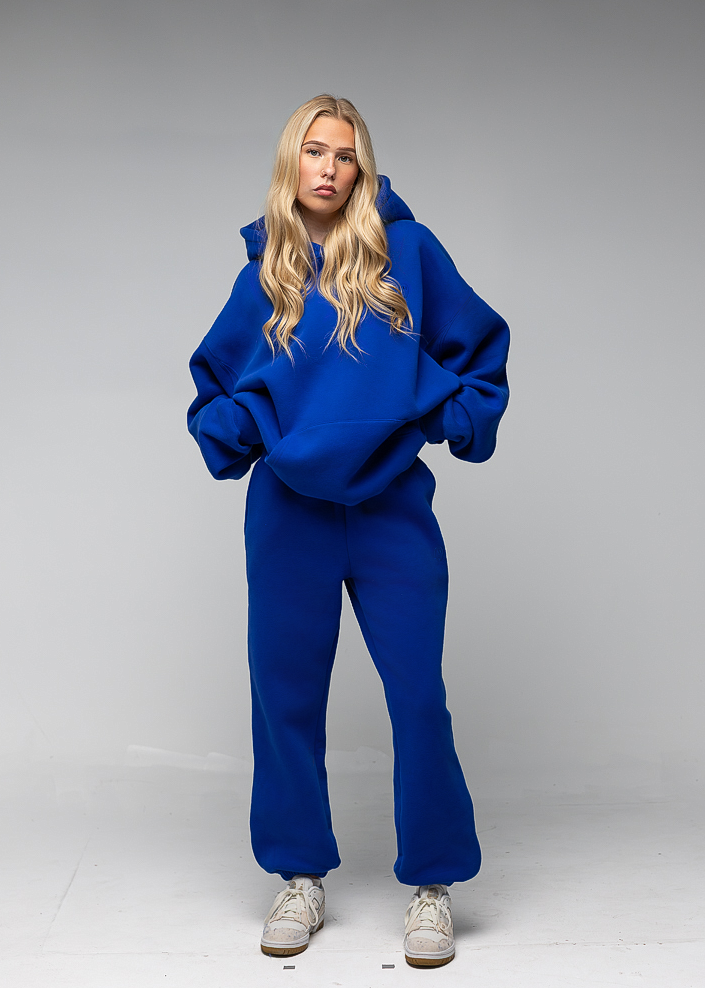 A blonde model wearing a matching royal blue sweatshirt and sweatpants set, paired with light gray sneakers, standing against a neutral gray studio background.