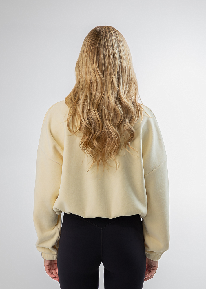 A woman with long blonde hair is shown from behind, wearing a cream-colored cropped sweatshirt and black leggings, standing against a light gray studio background.
