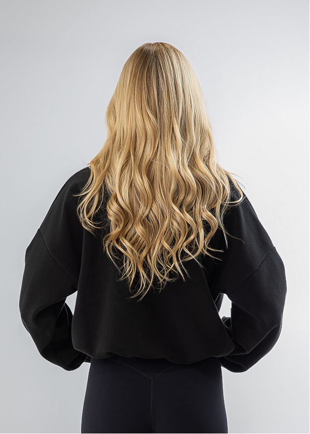 A woman with long blonde hair is shown from behind, wearing a black sweatshirt and black leggings with white sneakers, standing against a light gray studio background.