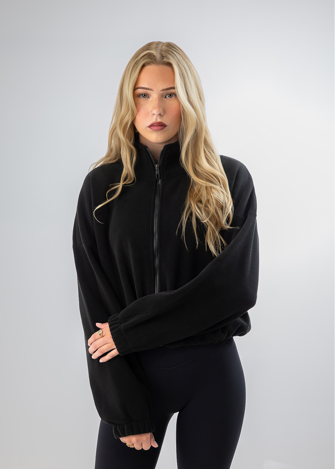 A blonde woman modeling a black zip-up fleece jacket with wide, balloon sleeves and black leggings, standing against a light gray studio background.