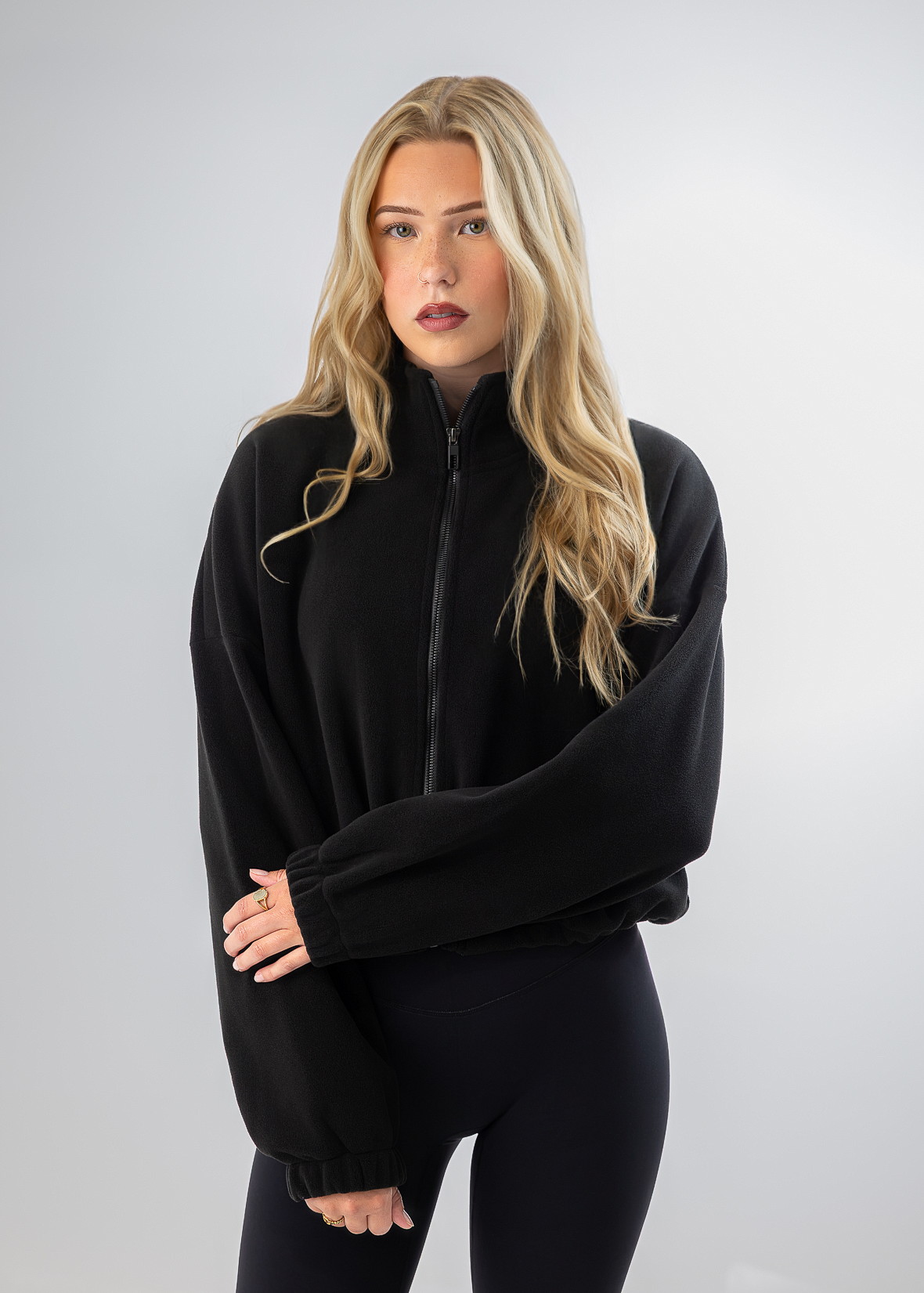A blonde woman modeling a black zip-up fleece jacket with long sleeves and black leggings against a light gray studio background.