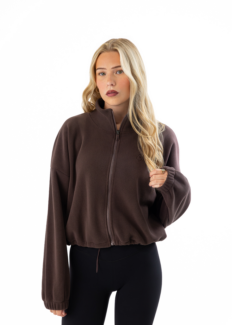 A blonde woman wearing a dark brown zip-up fleece jacket and black leggings, standing against a solid white background.