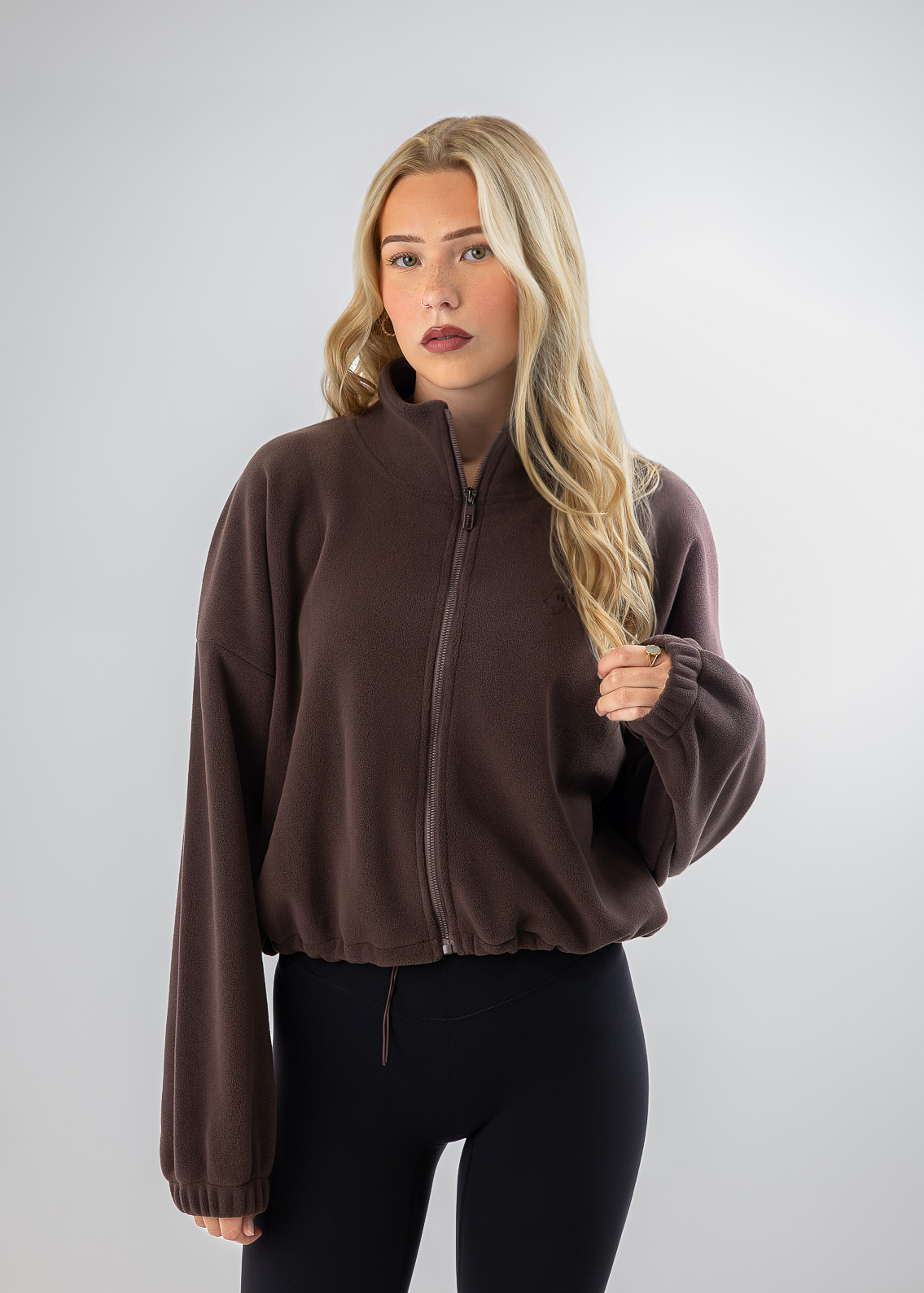 A blonde woman modeling a dark brown cropped zip-up fleece jacket with long sleeves and black leggings, standing against a light gray studio background.
