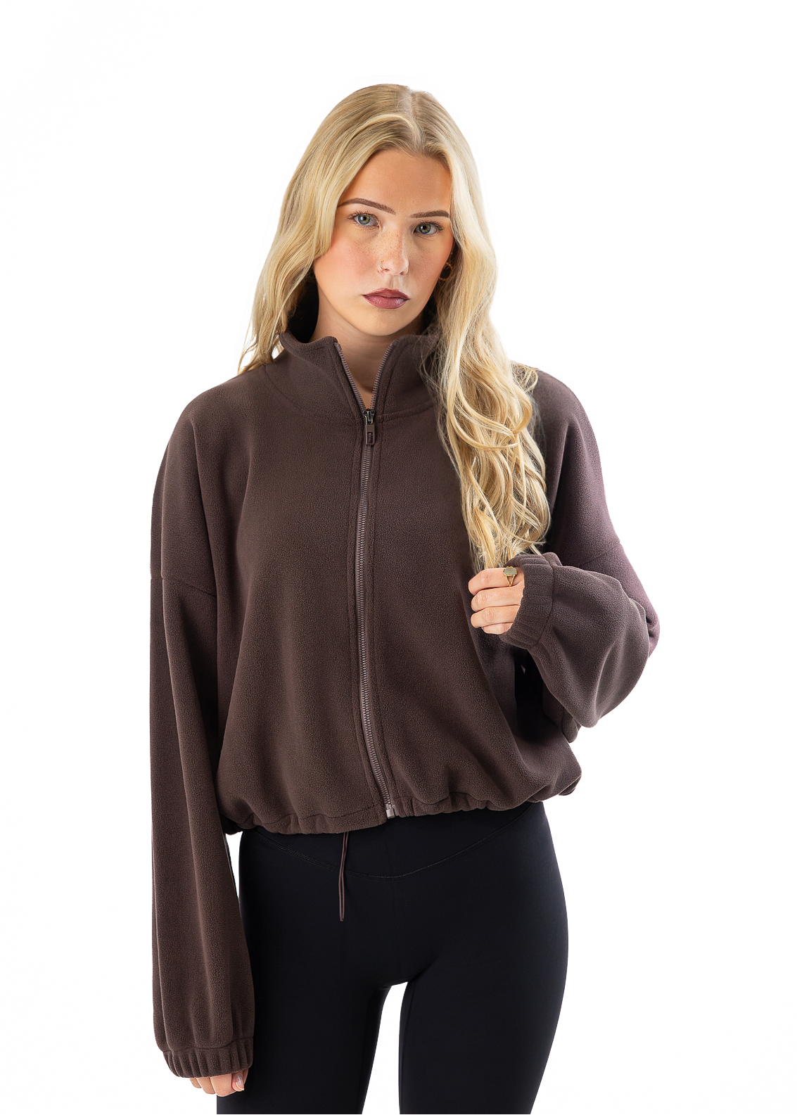 A blonde woman modeling a dark brown zip-up fleece jacket with a stand collar and long sleeves, paired with black leggings, against a solid white background.