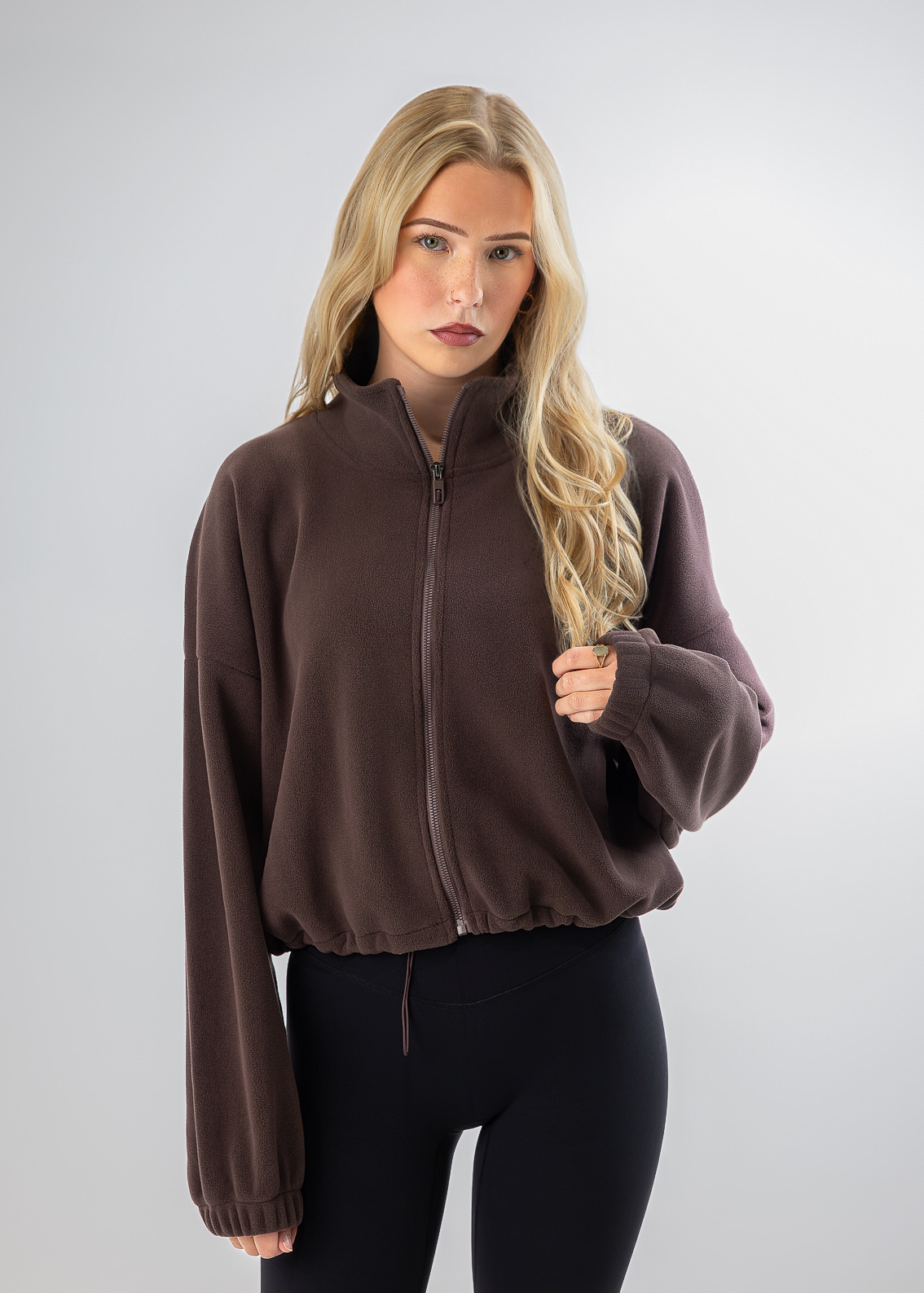 A blonde woman modeling a dark brown zip-up fleece jacket with a stand collar and elastic cuffs, paired with black leggings, against a light gray studio background.