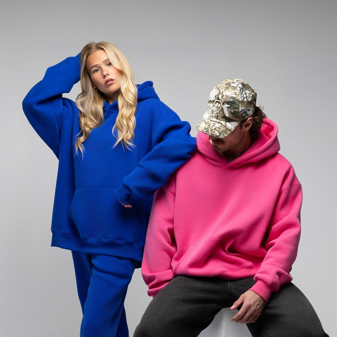 Two models posing in oversized streetwear clothing against a light gray studio background. One model wears a bright blue hoodie and matching sweatpants, while the other wears a vibrant pink hoodie, black sweatpants, a camouflage baseball cap, and black-and-white sneakers, sitting on a white geometric block.