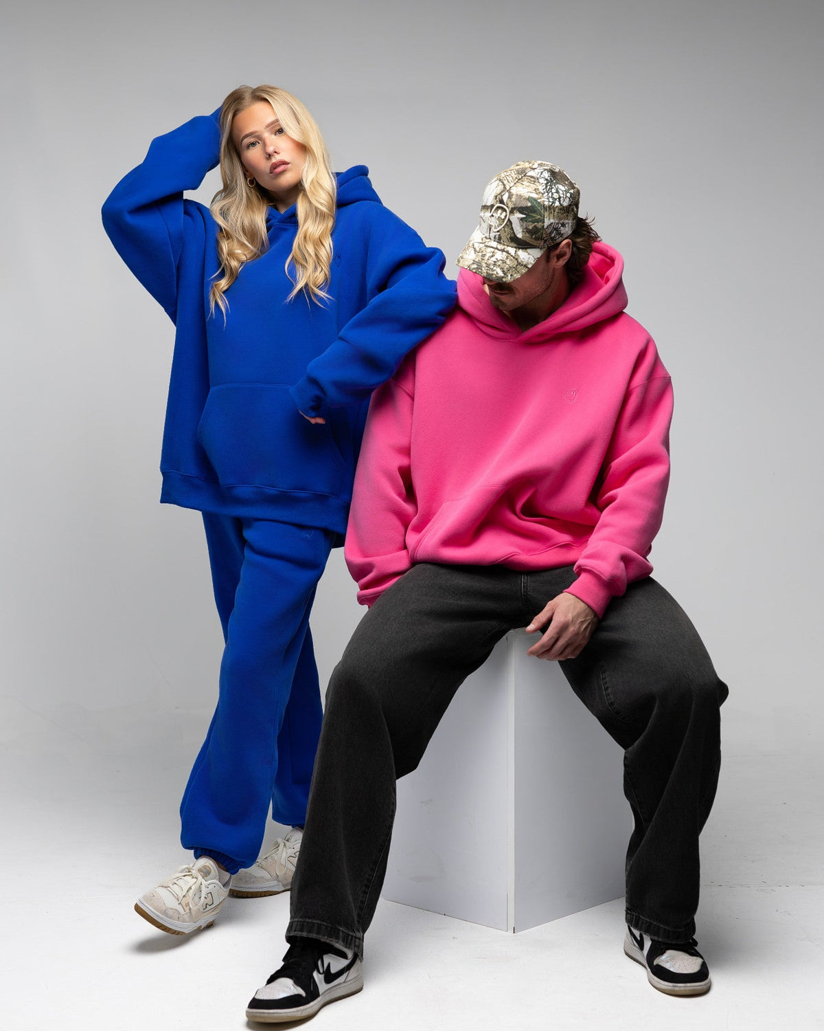 Two models posing in oversized streetwear clothing against a light gray studio background. One model wears a bright blue hoodie and matching sweatpants, while the other wears a vibrant pink hoodie, black sweatpants, a camouflage baseball cap, and black-and-white sneakers, sitting on a white geometric block.
