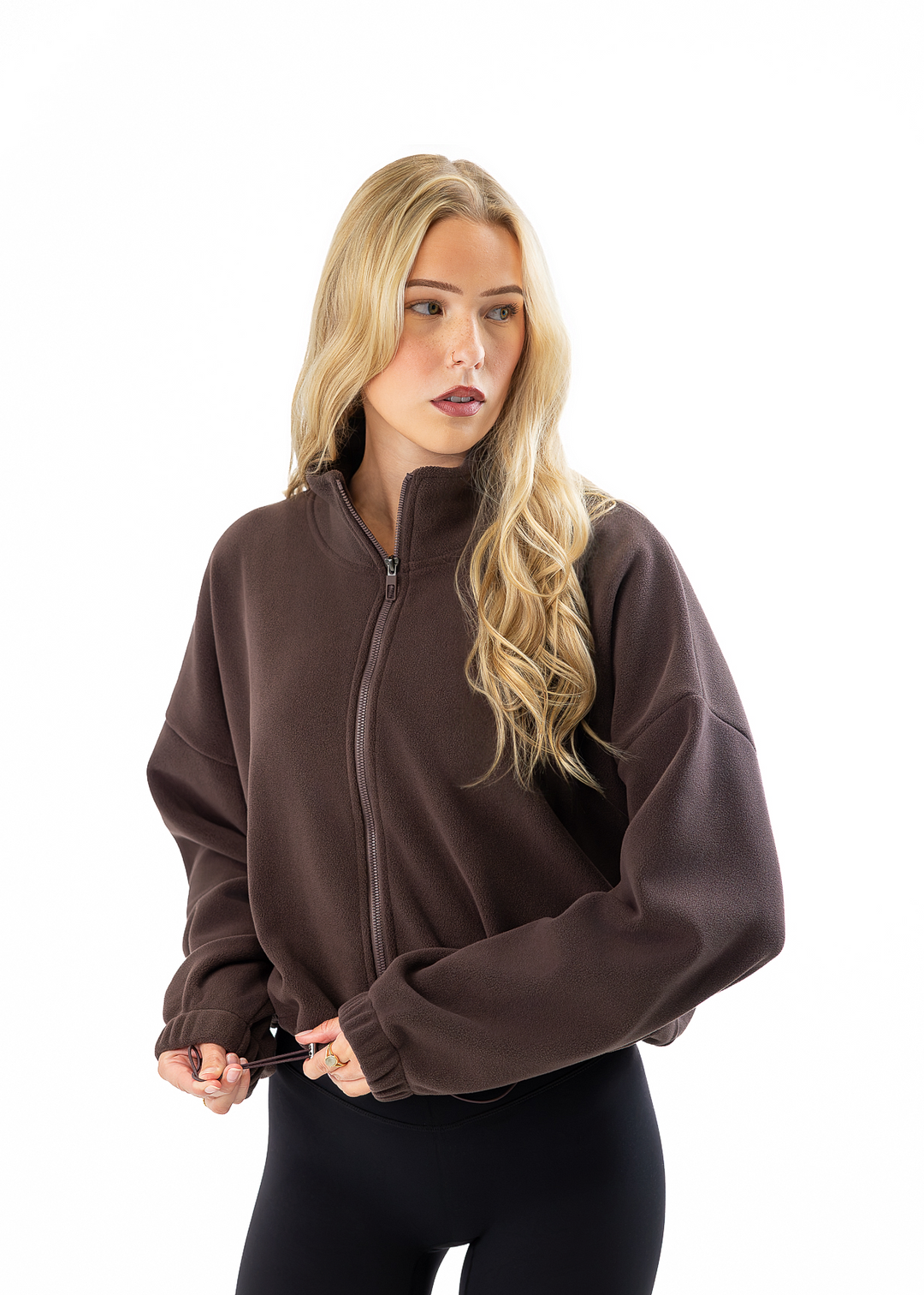 A blonde woman wearing a dark brown zip-up fleece jacket and black leggings, standing against a solid white background.