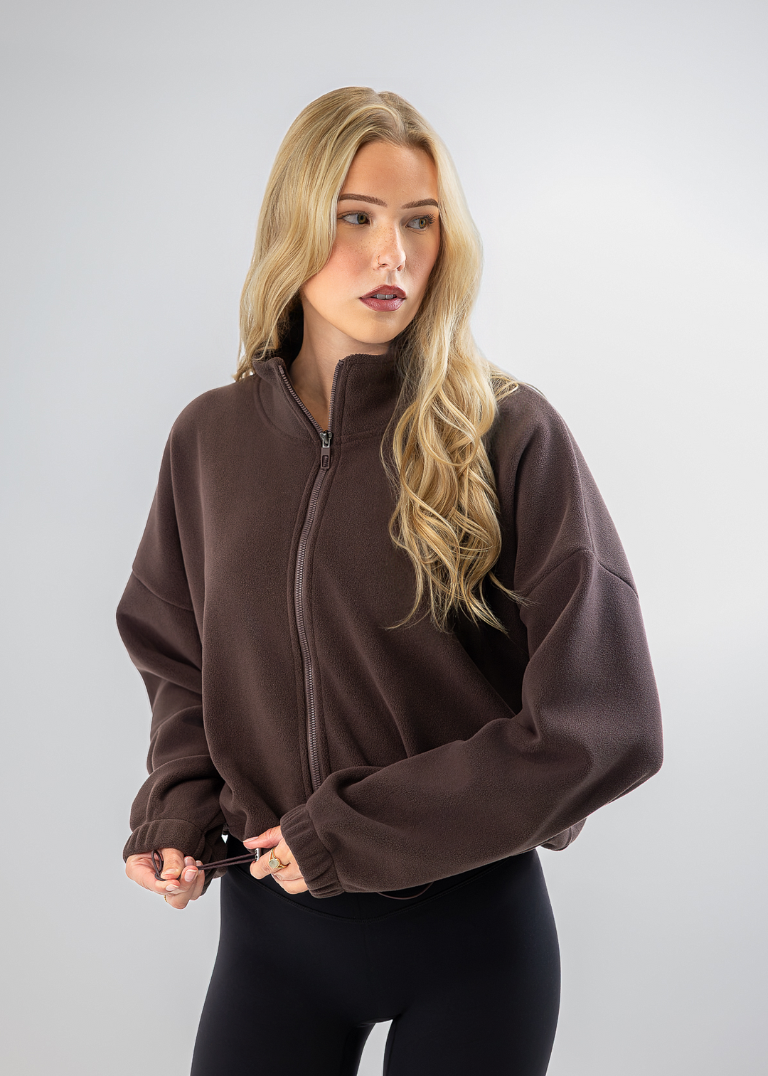 A blonde woman wearing a dark brown cropped zip-up fleece jacket and black leggings, standing against a light gray studio background.