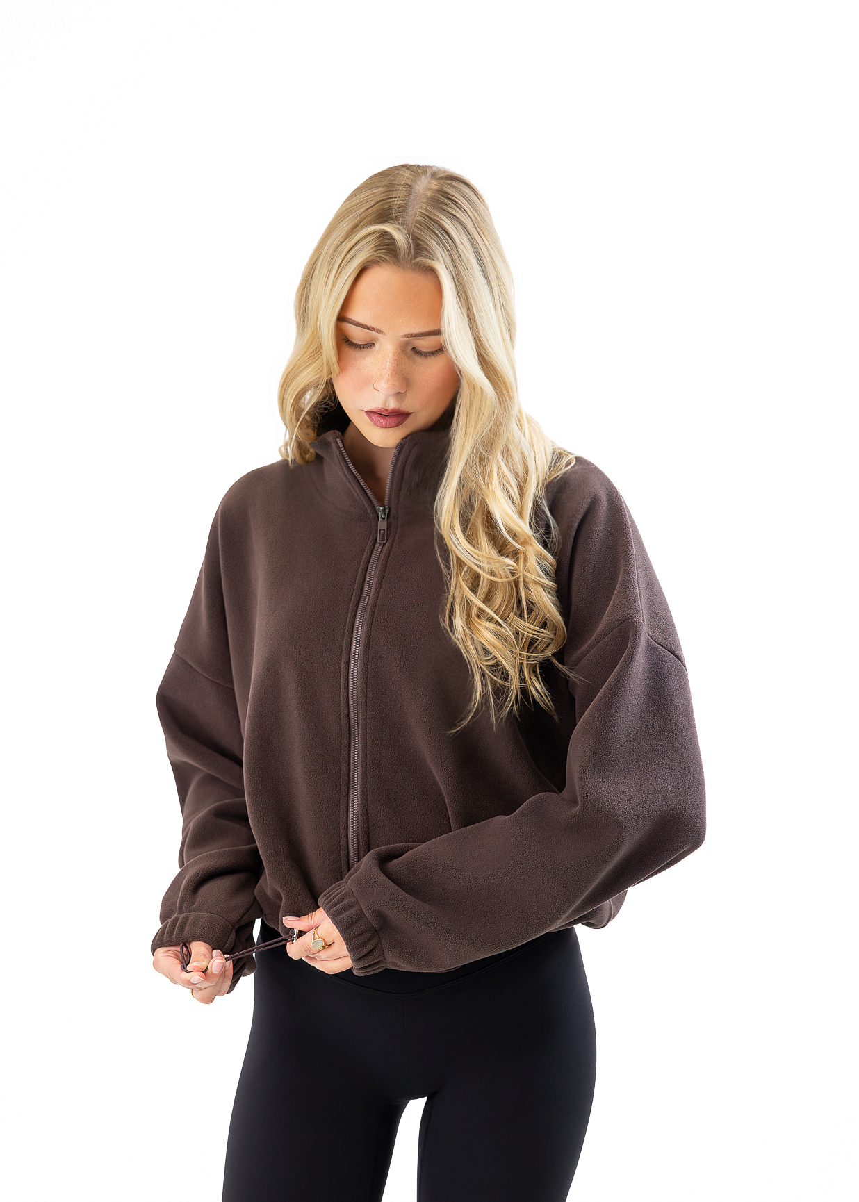 A blonde woman wearing a dark brown cropped zip-up fleece jacket and black leggings, standing against a solid white background.