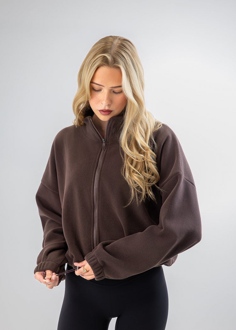 A blonde woman wearing a dark brown cropped zip-up fleece jacket and black leggings, standing against a light gray studio background.