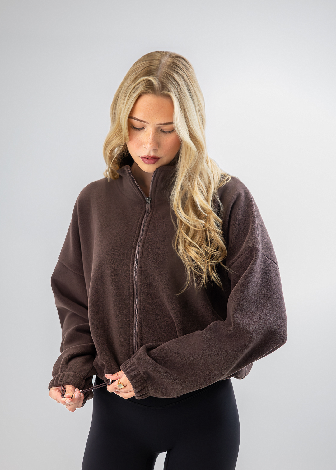 A blonde woman wearing a dark brown cropped zip-up fleece jacket and black leggings, standing against a light gray studio background.