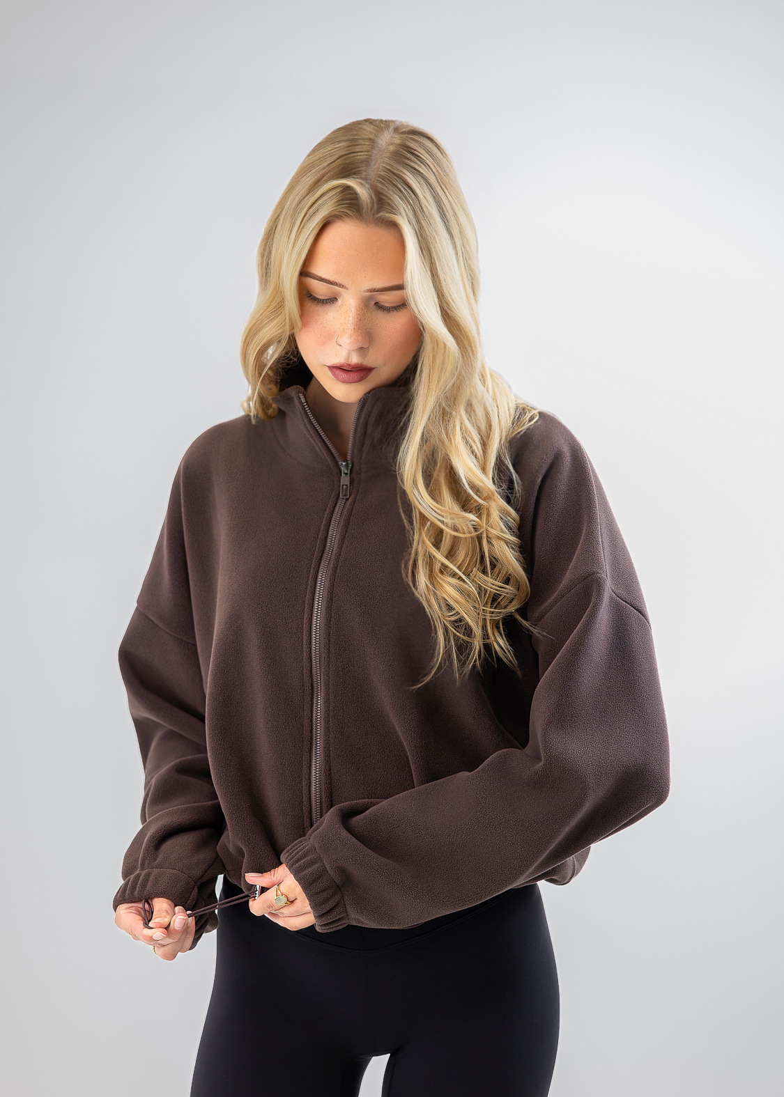 A blonde woman wearing a dark brown cropped zip-up fleece jacket and black leggings, standing against a light gray studio background.
