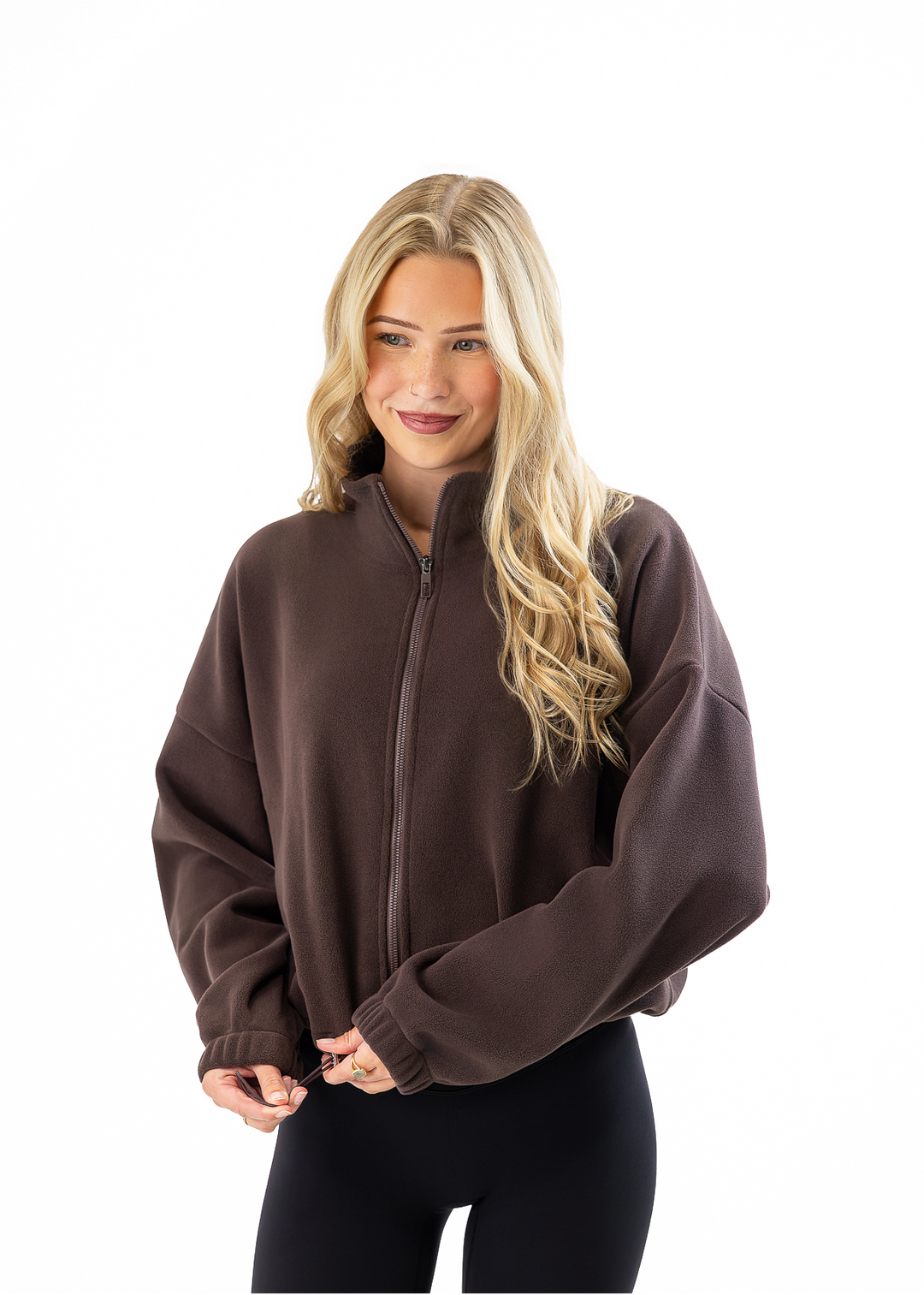 A woman with blonde hair wearing a dark brown cropped zip-up fleece jacket and black leggings, standing against a solid white background.