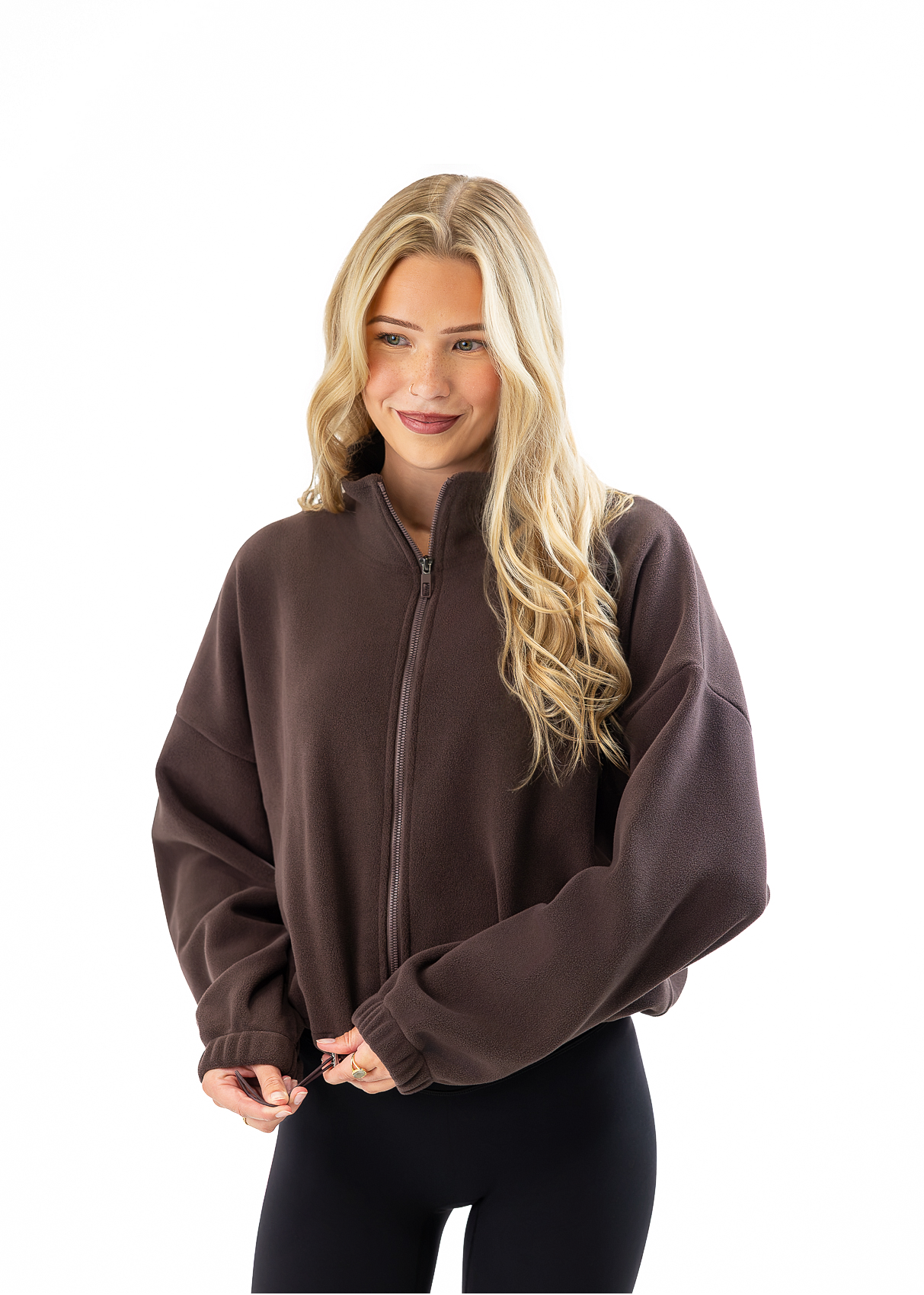 A woman with blonde hair wearing a dark brown cropped zip-up fleece jacket and black leggings, standing against a solid white background.