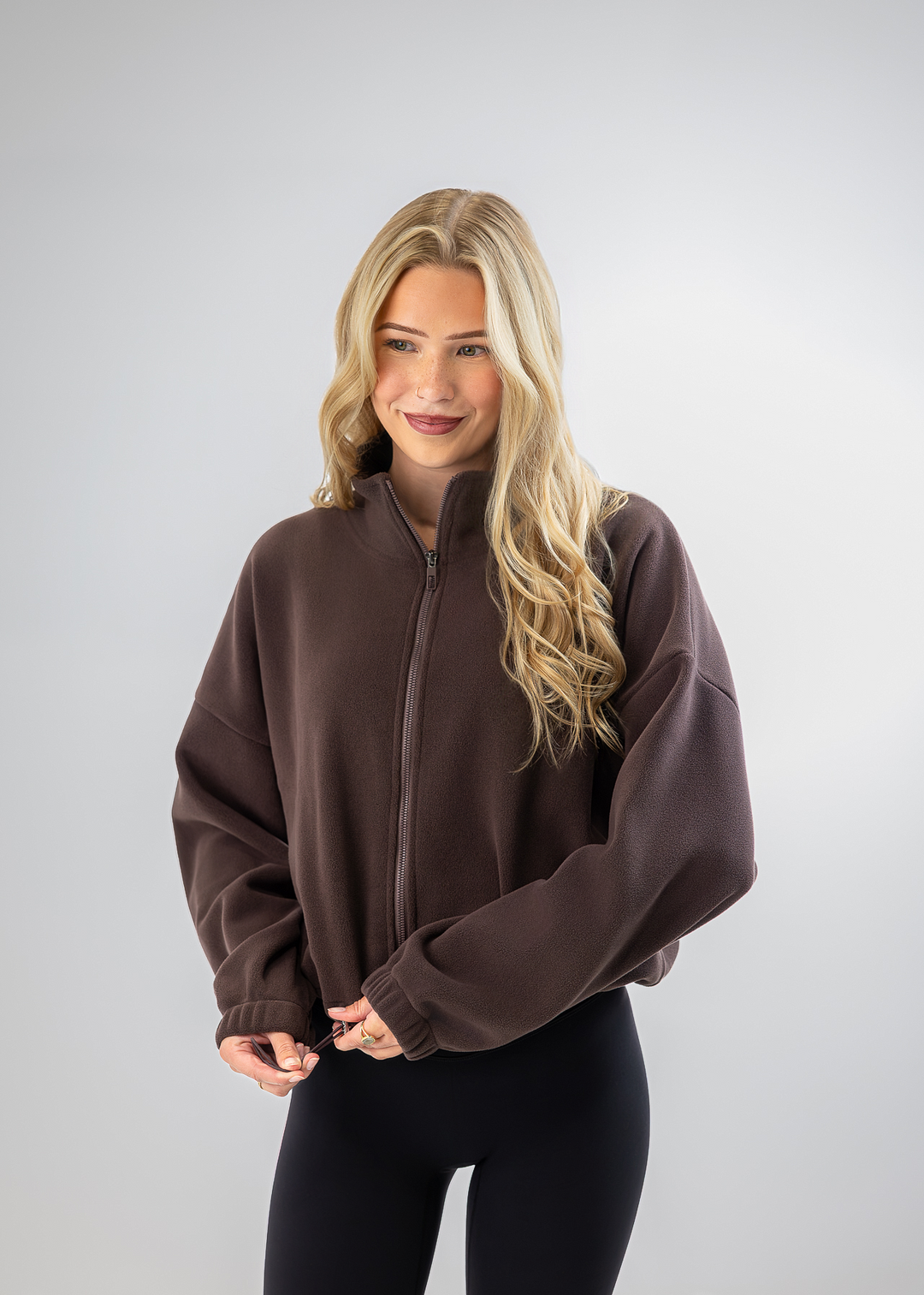 A woman with long blonde hair wearing a dark brown cropped zip-up fleece jacket and black leggings, standing against a light gray studio background.