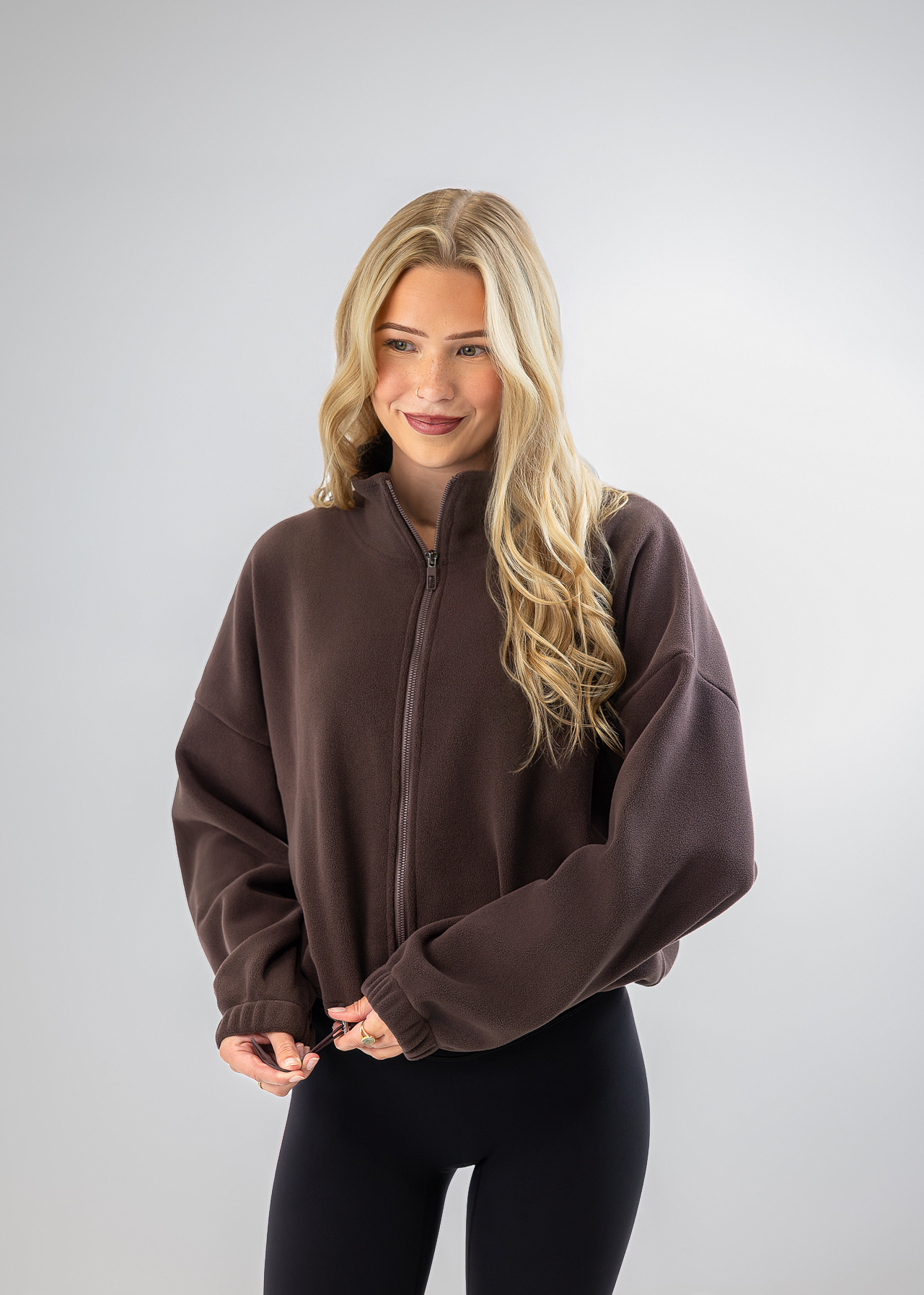 A woman with long blonde hair wearing a dark brown cropped zip-up fleece jacket and black leggings, standing against a light gray studio background.