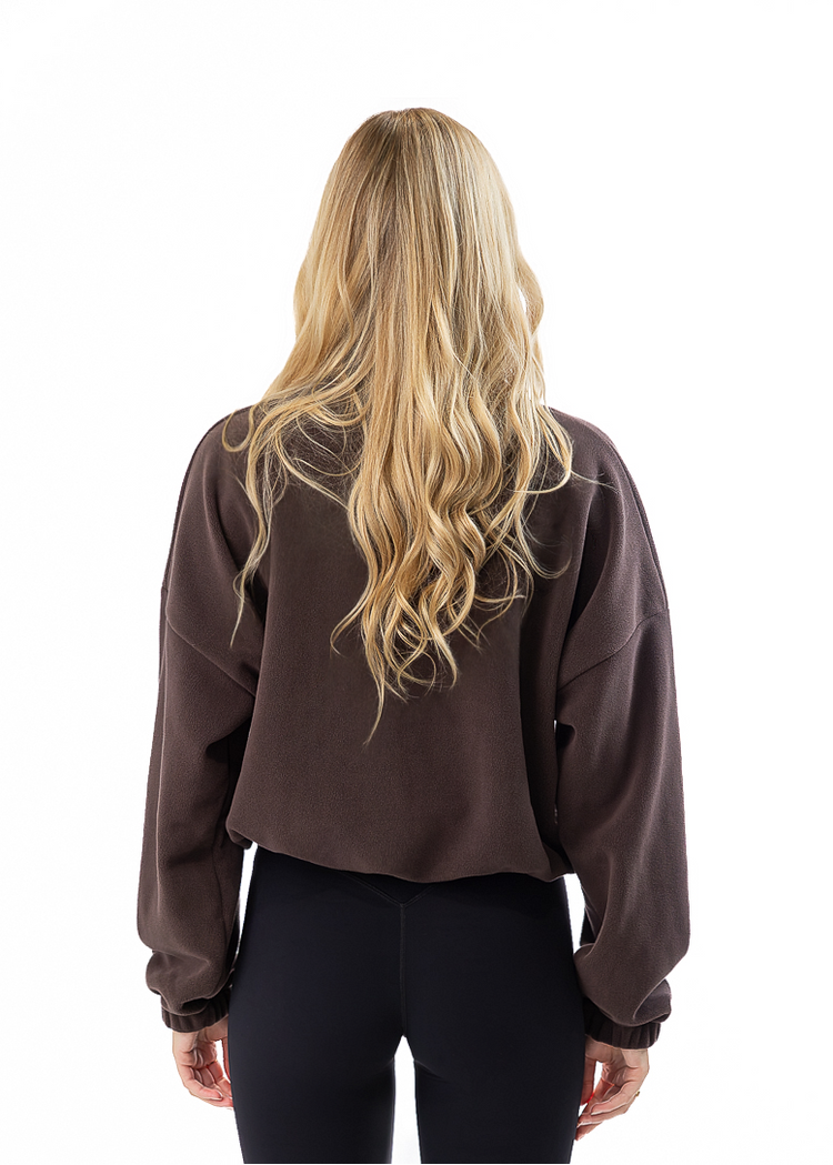 A woman with long blonde hair is shown from behind wearing a dark brown cropped sweatshirt, black leggings, and white sneakers against a solid white background.