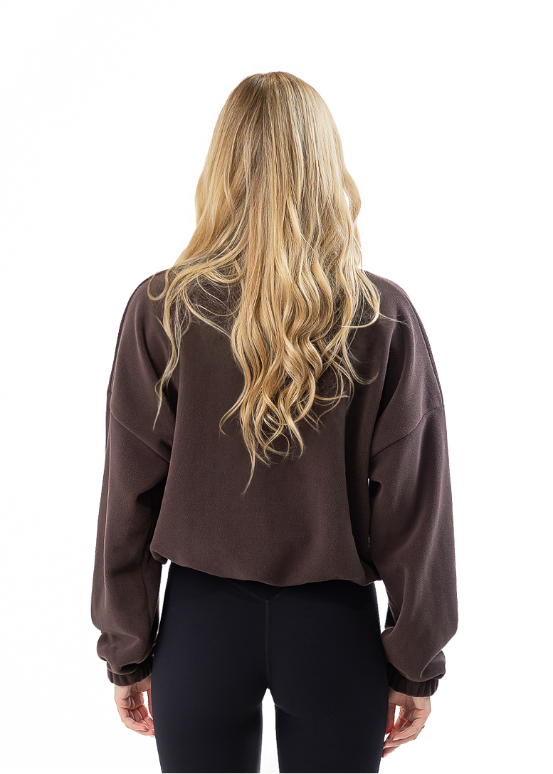 A woman with long blonde hair is shown from behind wearing a dark brown cropped sweatshirt, black leggings, and white sneakers against a solid white background.