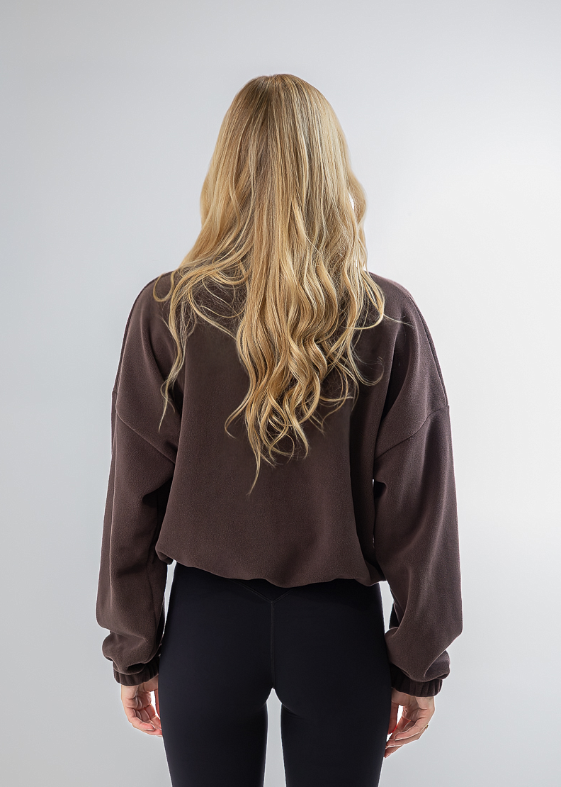 A woman with long blonde hair is shown from behind wearing a dark brown cropped sweatshirt, black leggings, and off-white sneakers against a light gray studio background.