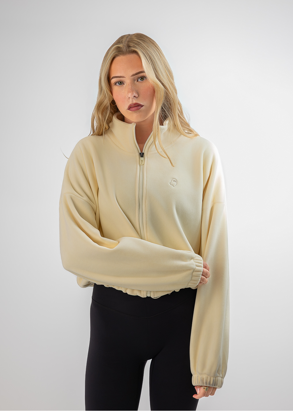 A blonde model wearing a cream-colored quarter-zip sweatshirt and black leggings, standing against a light gray studio background.