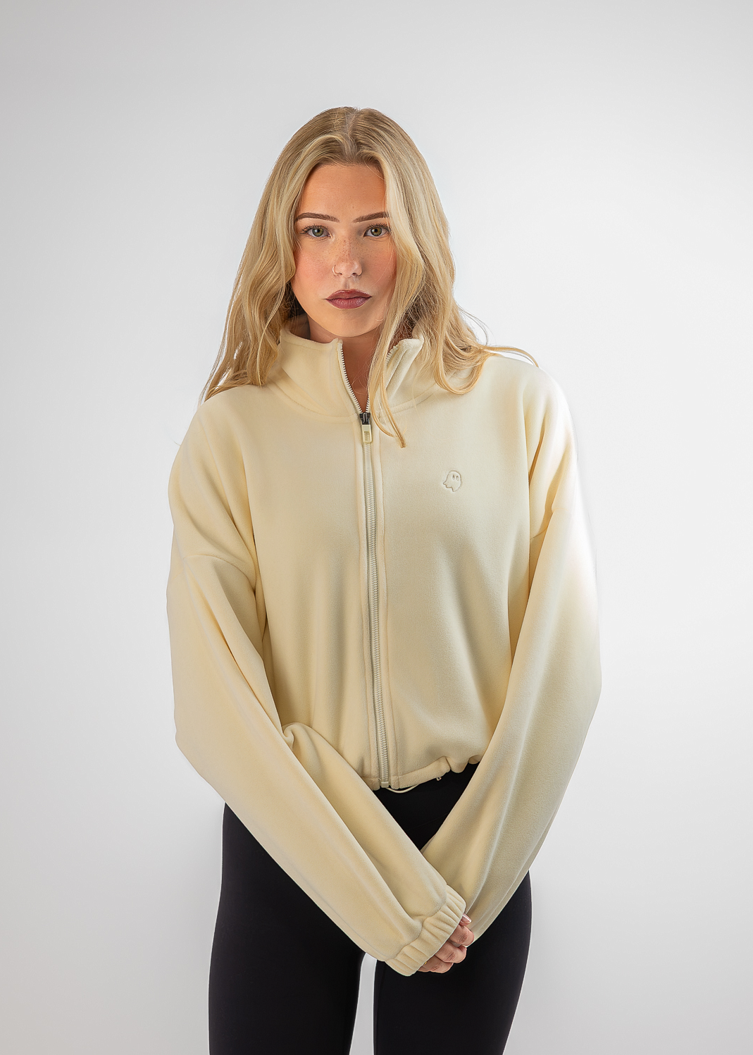 A blonde woman modeling a cream-colored zip-up hoodie with long sleeves and black leggings, standing against a light gray studio background.