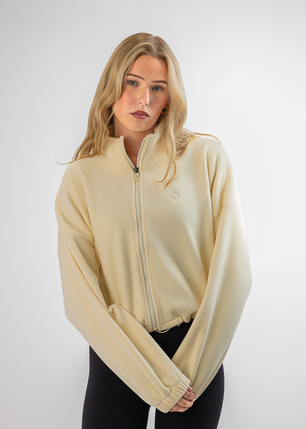 A blonde woman modeling a cream-colored zip-up fleece jacket with long sleeves and black leggings, standing against a light gray studio background.
