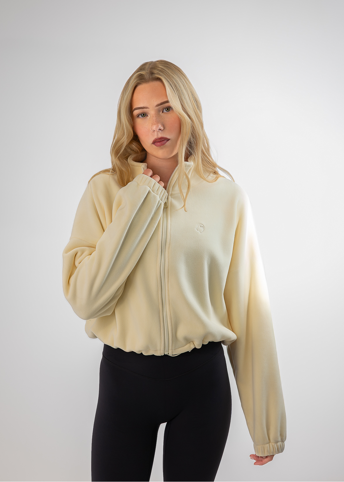 A blonde woman modeling a cream-colored zip-up fleece jacket with black leggings, standing against a light gray studio background.