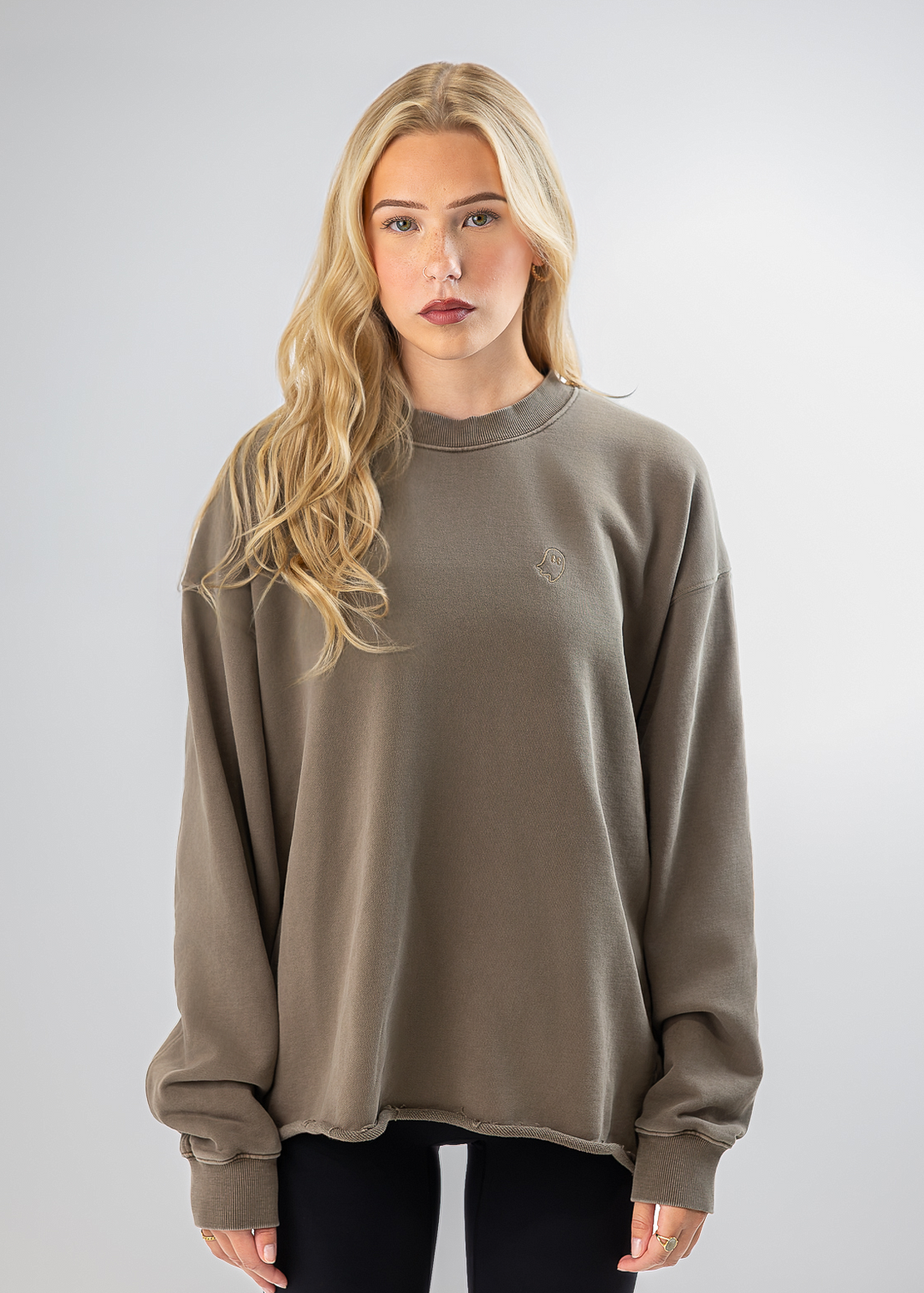 A blonde woman modeling an oversized crewneck sweatshirt in a muted taupe-brown color, paired with black pants, against a light gray studio background.