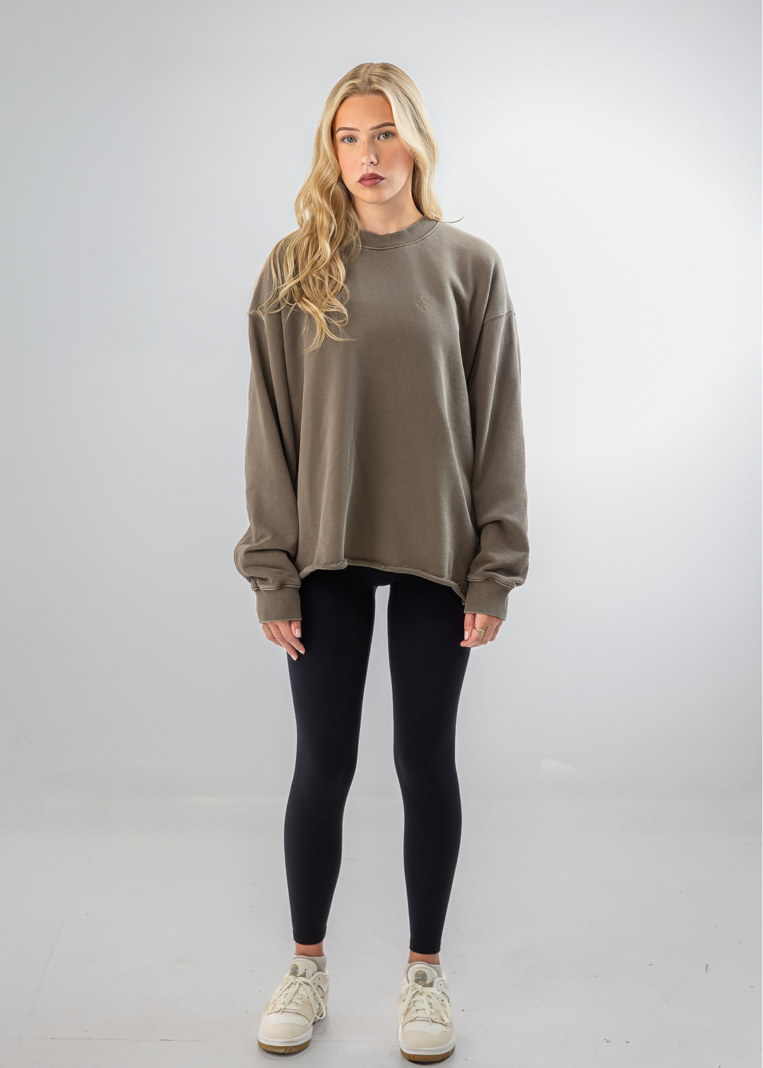 A blonde woman stands against a light gray studio background wearing an oversized taupe crewneck sweatshirt, black leggings, and off-white sneakers.