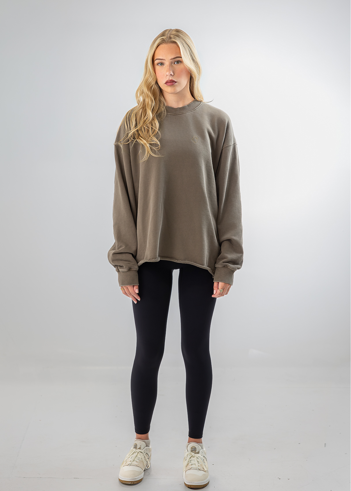 A blonde woman stands against a light gray studio background wearing an oversized taupe crewneck sweatshirt, black leggings, and off-white sneakers.