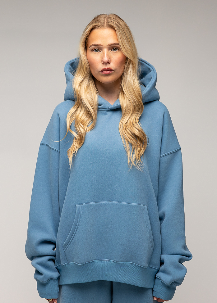 A blonde woman wearing an oversized dusty blue hoodie and matching sweatpants, standing against a neutral gray studio background.