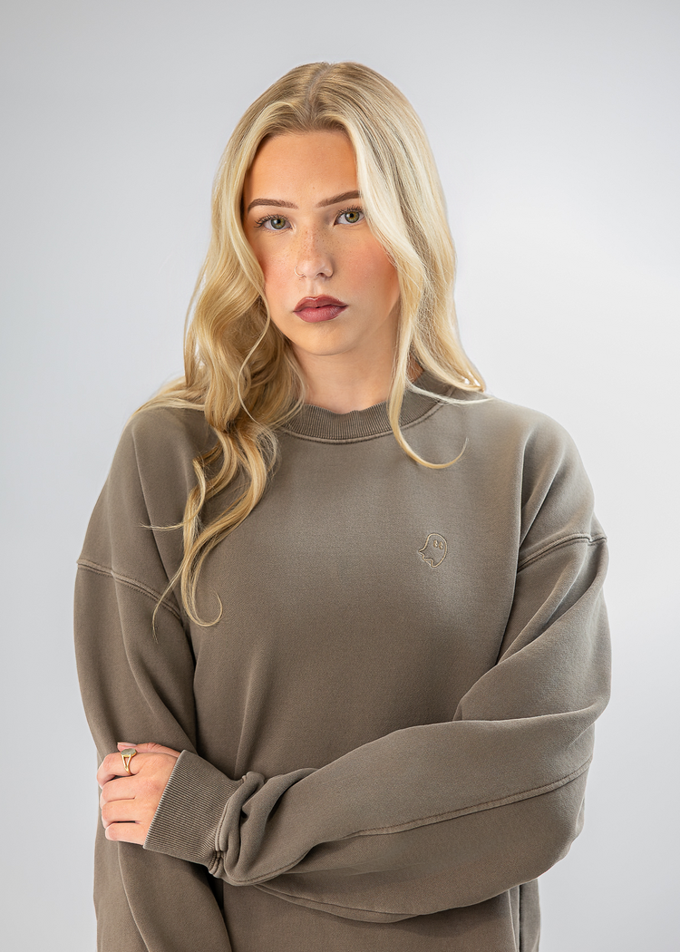 A blonde woman wearing an oversized taupe crewneck sweatshirt, standing against a light gray studio background with her arms crossed.