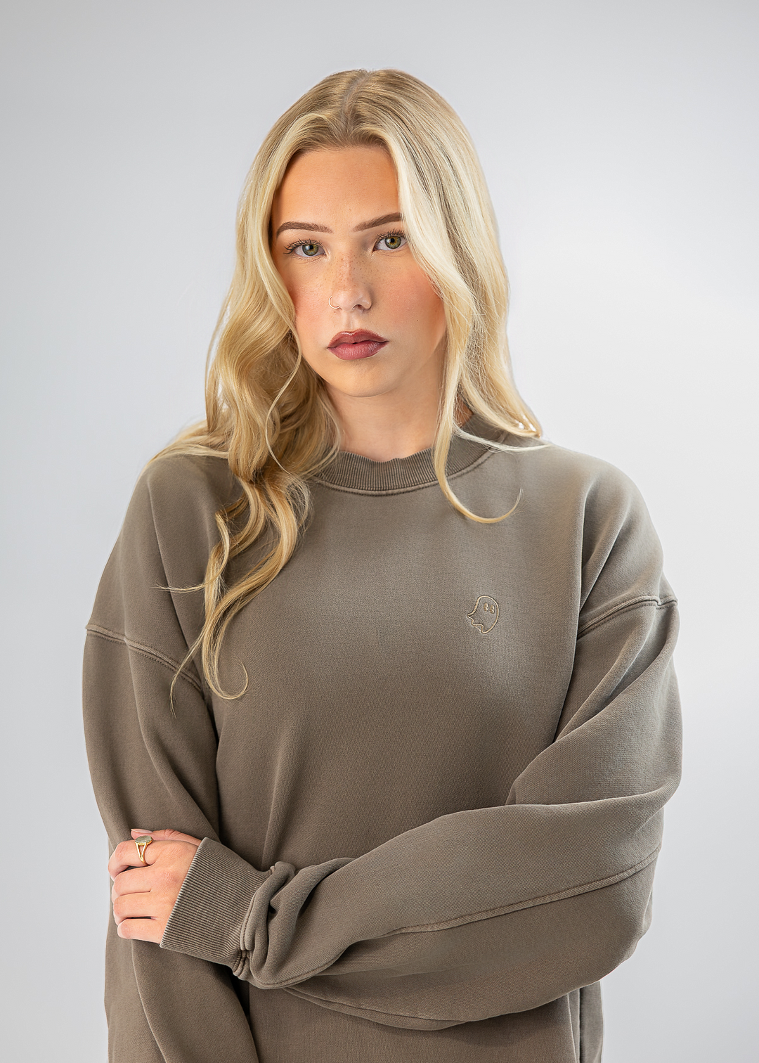 A blonde woman wearing an oversized taupe crewneck sweatshirt, standing against a light gray studio background with her arms crossed.
