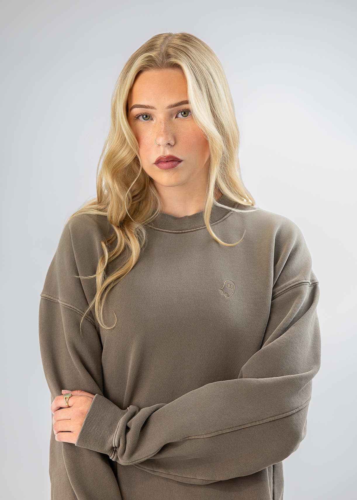 A blonde woman wearing an oversized taupe crewneck sweatshirt, standing against a light gray studio background with her arms crossed.