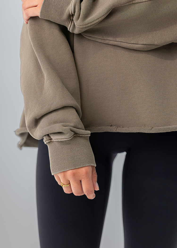 A close-up of a person wearing an oversized taupe sweatshirt with rolled cuffs and black leggings, cropped from the chest down against a light gray studio background.