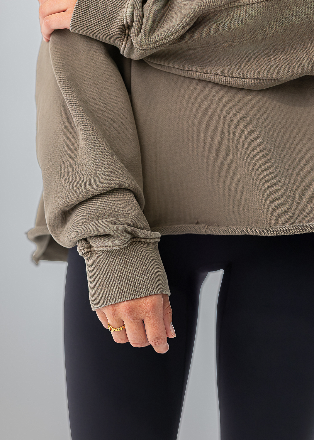 A close-up of a person wearing an oversized taupe sweatshirt with rolled cuffs and black leggings, cropped from the chest down against a light gray studio background.