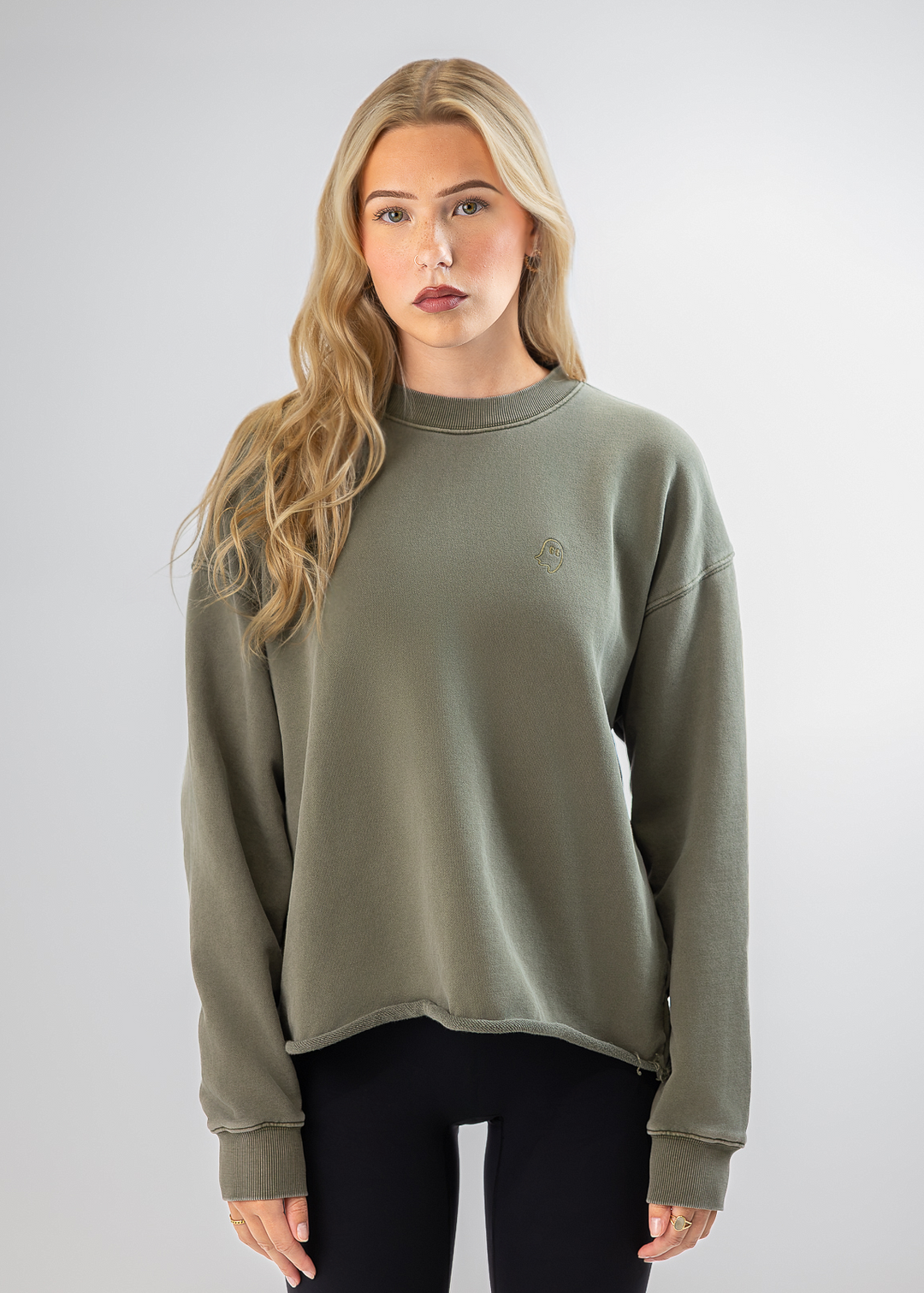 A blonde woman modeling a muted olive green crewneck sweatshirt with a small embroidered logo on the left chest, paired with black leggings, against a light gray studio background.