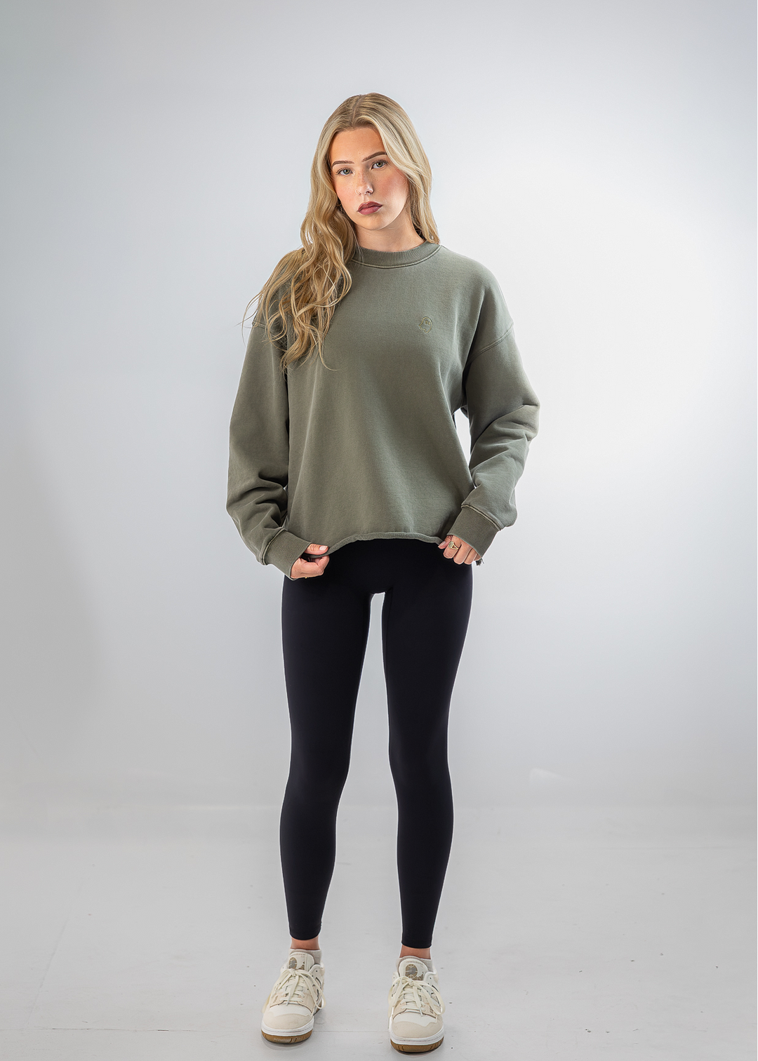A woman modeling a sage green crewneck sweatshirt, black leggings, and white sneakers against a light gray studio background.