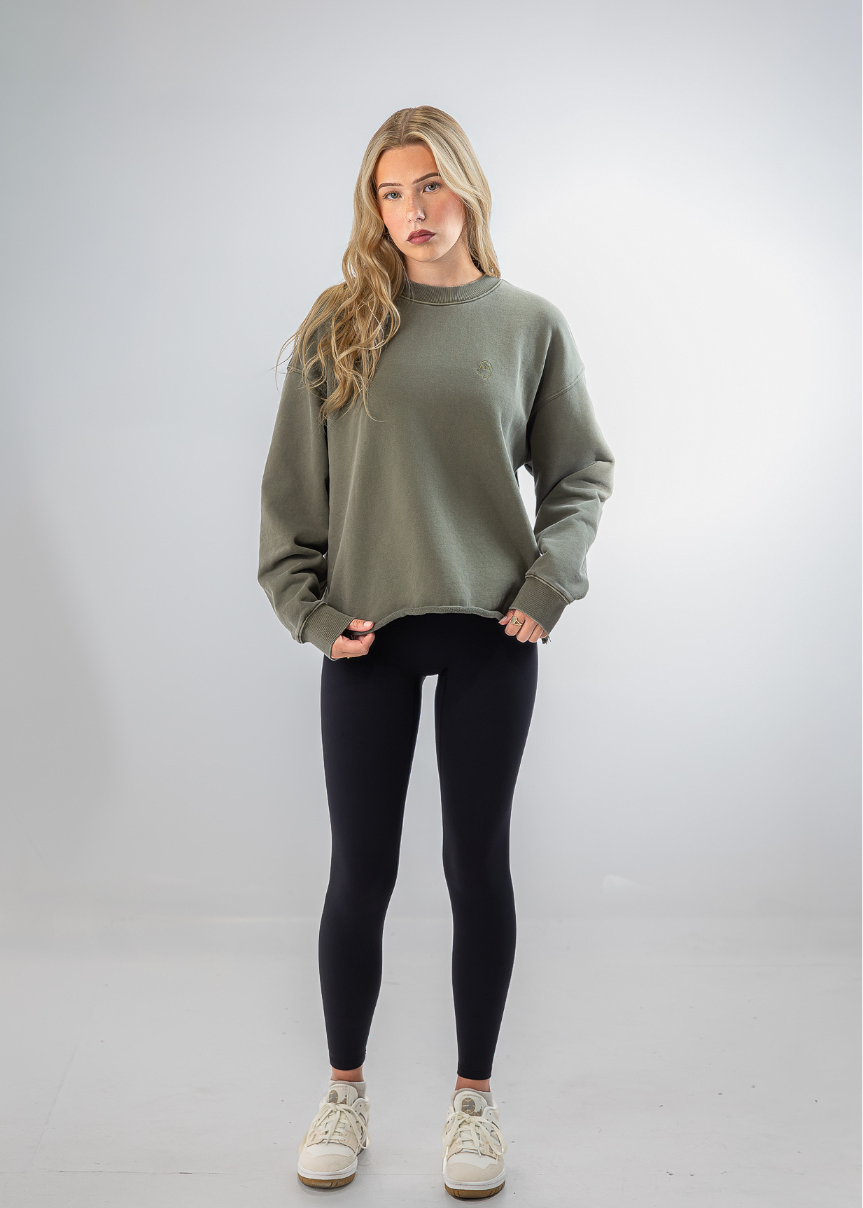 A woman modeling a sage green crewneck sweatshirt, black leggings, and white sneakers against a light gray studio background.