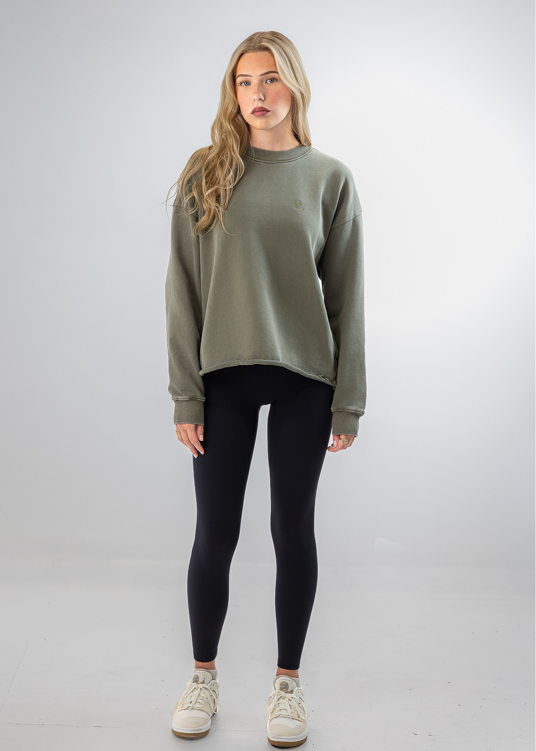 A woman modeling a loose-fit sage green crewneck sweatshirt, paired with black leggings and off-white sneakers, standing against a light gray studio background.