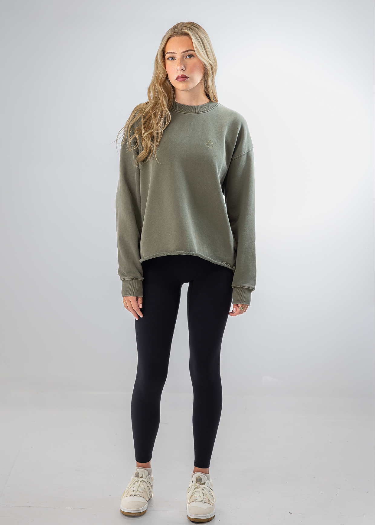 A woman modeling a loose-fit sage green crewneck sweatshirt, paired with black leggings and off-white sneakers, standing against a light gray studio background.