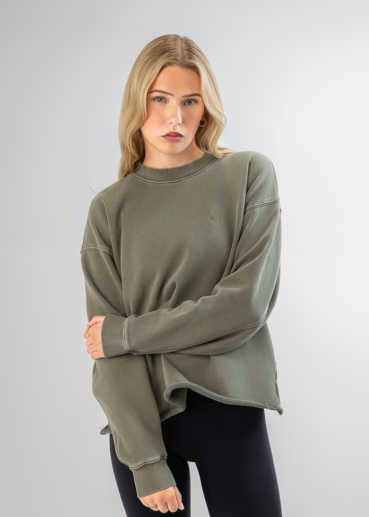 A woman modeling an oversized crewneck sweatshirt in olive green, paired with black leggings, against a light gray studio background.
