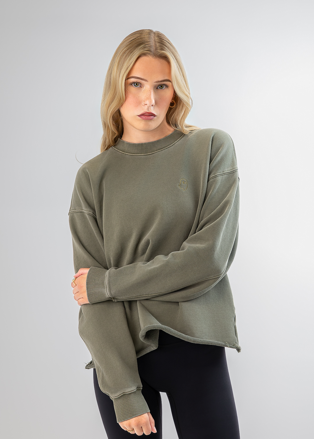 A woman modeling an oversized crewneck sweatshirt in olive green, paired with black leggings, against a light gray studio background.